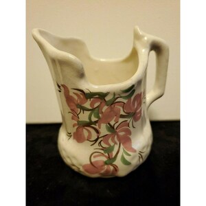 May include: A small white ceramic pitcher with a floral design. The pitcher has a handle and a spout. The flowers are pink and green.