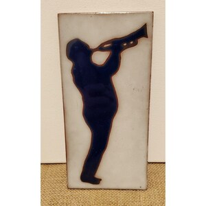 Puede incluir: Un azulejo de cerámica blanco con una silueta azul de una persona tocando la trompeta. El azulejo tiene un aspecto rústico y pintado a mano.