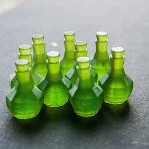 May include: A group of ten small, green glass bottles with a faceted design. The bottles are arranged in a semi-circle on a gray surface.