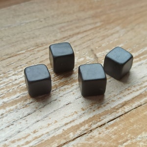 Op de afbeelding: Vier kleine, donkergrijze vierkante objecten zijn gerangschikt op een lichtbruin houten oppervlak. De objecten lijken op dobbelstenen of vergelijkbare spelstukken, elk met een gladde, matte afwerking. De houtnerf zorgt voor een natuurlijke achtergrond.