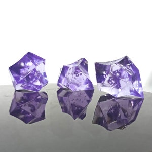 Op de afbeelding: Drie heldere paarse glazen decoratieve edelstenen met een facetgeslepen ontwerp. De edelstenen zijn in een rij op een reflecterend oppervlak gerangschikt.