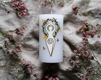 Könnte beinhalten: Weiße Kerze mit einem goldenen und grünen bemalten Design einer Frau mit einem Kranz aus Blumen um den Kopf. Die Kerze ist von kleinen rosa und weißen Blumen umgeben.