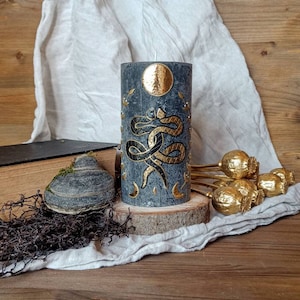 Peut inclure: Une bougie grise avec un motif de serpent doré et des phases de lune. La bougie est sur une base en bois et entourée de capsules de pavot dorées.