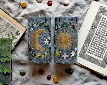 May include: Two gray pillar candles with floral designs. One candle features a gold crescent moon and the other features a gold sun.
