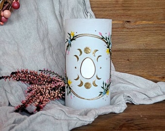 Könnte beinhalten: Eine weiße Kerze in Säulenform mit einem goldenen und weißen Mond- und Sonnenmotiv, umgeben von einem floralen Kranz mit rosa, gelben und blauen Blumen. Die Kerze steht auf einer hellgrauen Stoffoberfläche.