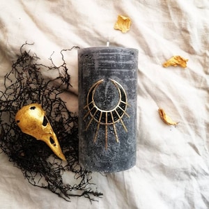 May include: A black candle with a gold geometric design on the side. The candle is sitting on a white fabric surface with a gold bird skull and dried seaweed.