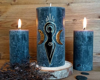 Könnte beinhalten: Drei schwarze Kerzen mit goldenen Akzenten. Die mittlere Kerze zeigt ein Dreifachmond-Symbol und eine stilisierte Frauenfigur. Die Kerzen stehen auf einer Holzoberfläche mit einem Stück Treibholz und schwarzen Steinen.