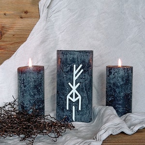 Könnte beinhalten: Drei schwarze Kerzen mit einem weißen Symbol auf der mittleren Kerze. Die Kerzen stehen auf einer weißen Stoffoberfläche mit einem Holz Hintergrund.