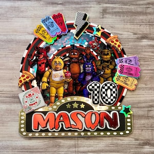 May include: A colorful cake topper featuring characters from the video game Five Nights at Freddy's. The topper includes the name "MASON" in red, the number "10", and various pizza and ticket-themed decorations. The characters are in various colors.
