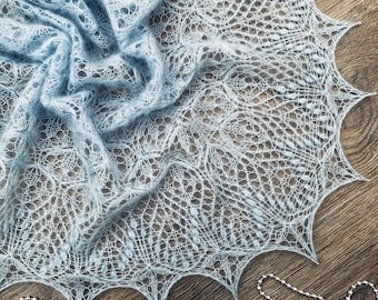 Pode incluir: Um xale delicado de cor azul claro, tricotado com um padrão de renda. O xale está drapeado sobre uma superfície de madeira e tem uma franja ao redor das bordas.