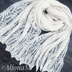 May include: A white crocheted shawl with a delicate lace pattern. The shawl is folded and draped over a dark surface. #AlionaM