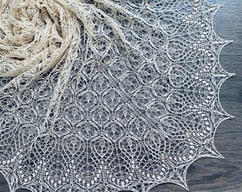 Pode incluir: Um xale de crochê delicado de cor branco-queimado com um padrão floral de renda. O xale está espalhado sobre uma superfície cinza escura.