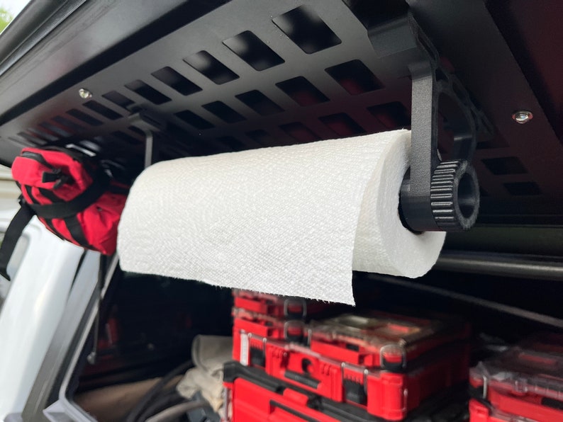 May include: A black metal paper towel holder mounted to a black metal grid. A white paper towel roll is attached to the holder. The holder has a knob to adjust the tension of the paper towel roll.