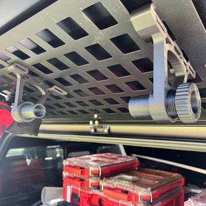 May include: Two black metal hooks with a threaded knob on the end are mounted to a black metal grid. The hooks are hanging from the ceiling of a vehicle.