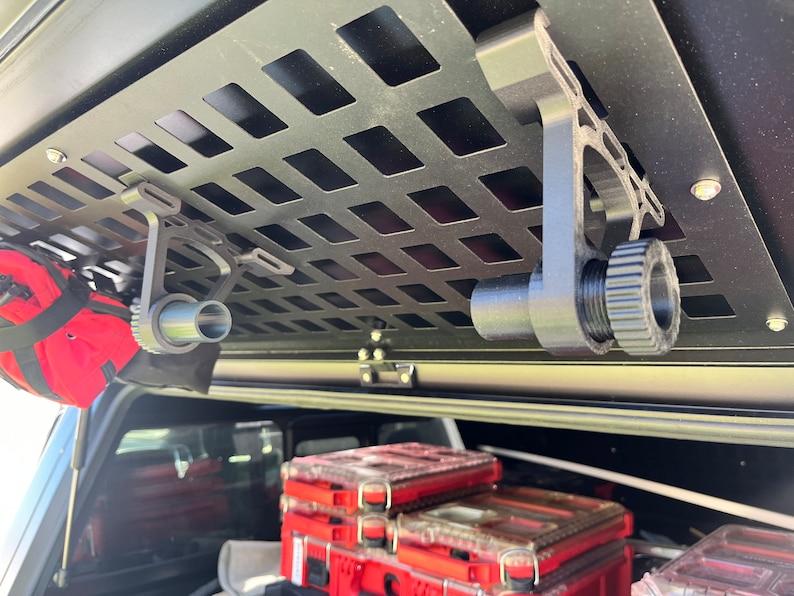 May include: Two black metal hooks with a threaded knob on the end are mounted to a black metal grid. The hooks are hanging from the ceiling of a vehicle.
