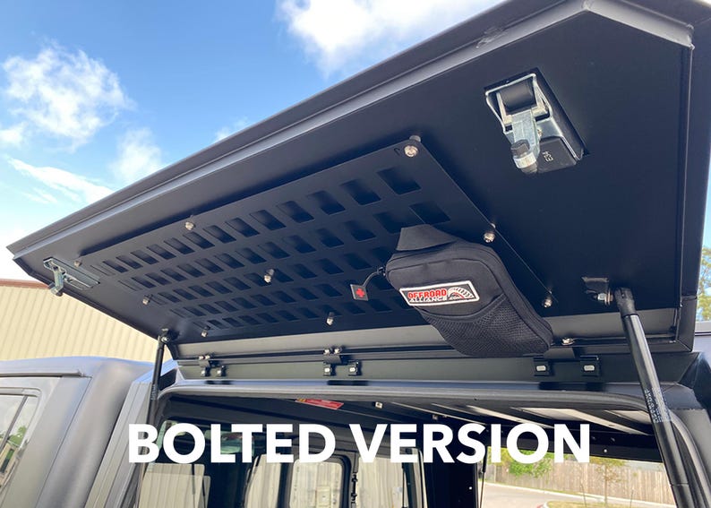 May include: A black metal roof rack with a bolted-on cargo shelf and a black mesh panel. The shelf has a small black pouch attached to it. The text "BOLTED VERSION" is visible in the image.