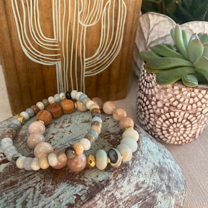 May include: Two beaded bracelets with a mix of light blue, brown, and pink stones. The bracelets are stacked on top of each other on a blue and brown wooden surface.