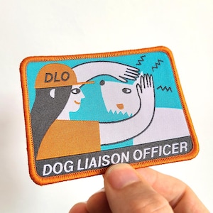 May include: Embroidered patch with a teal and white background, featuring a cartoon illustration of a person and a dog. The person wears an orange cap with "DLO" on it. The text "DOG LIAISON OFFICER" is at the bottom, with an orange border.