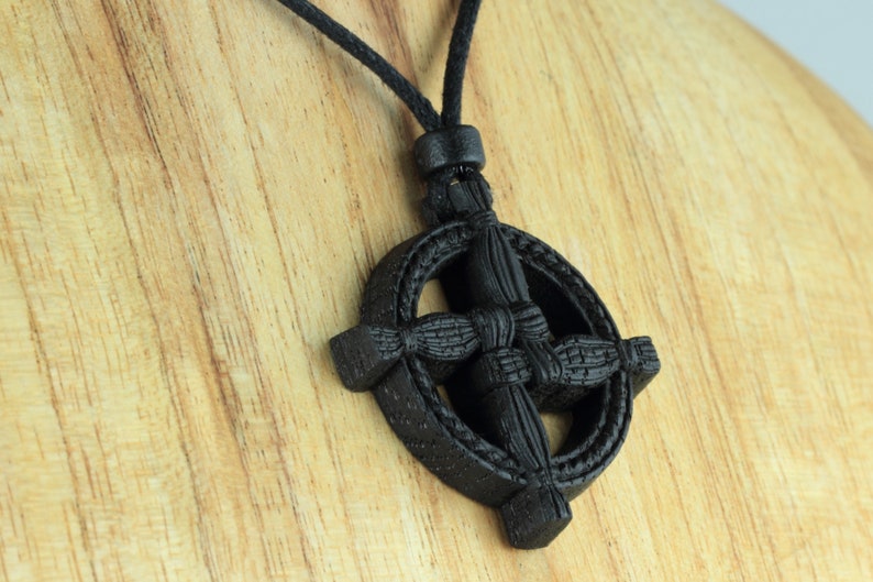 Puede incluir: Un colgante de madera negra con nudo celta en forma de cruz, en un collar de cord&oacute;n negro. El colgante est&aacute; tallado con un dise&ntilde;o de nudo celta.