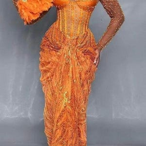 May include: A woman wearing a long, orange, beaded gown with a matching head wrap and holding a large orange feather fan.