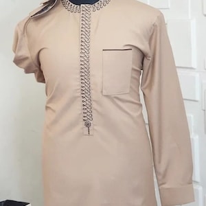 May include: A beige long-sleeved shirt with a decorative black trim around the collar and down the front. The shirt has a single button closure and a pocket on the left side.