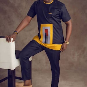 May include: A man wearing a black short-sleeved shirt with a yellow, grey, and red geometric design on the front. He is also wearing matching black trousers and brown loafers.