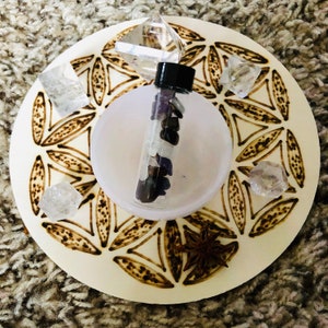 May include: A white wooden tray with a flower of life design burned into the surface. The tray has a small white bowl and a glass vial filled with various crystals. The vial has a black lid. There is a star anise on the tray.