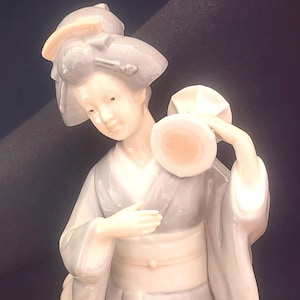 May include: A porcelain figurine of a woman in a traditional Japanese kimono, holding a small drum. The figurine is painted in shades of white, gray, and pink.