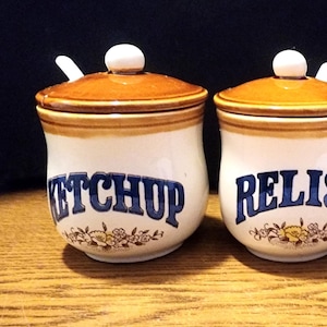Może przedstawiać: Trzy ceramiczne słoiki na przyprawy z brązowymi pokrywkami i białymi korpusami. Słoiki są oznaczone napisami "Ketchup", "Relish" i "Mustard" niebieskimi literami.