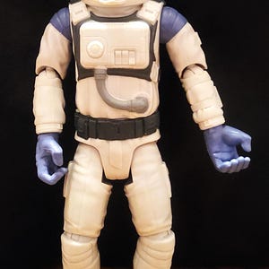 May include: A Buzz Lightyear action figure in a white and blue spacesuit. The figure has a blue helmet, gloves, and accents. The spacesuit has a white chest plate with a black belt and gray boots. The figure is standing upright.