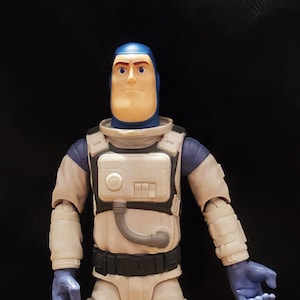 May include: A white and blue astronaut action figure with a blue helmet and a serious expression. The figure is wearing a white spacesuit with a black belt and a gray chest piece with a circular button and a hose.