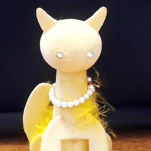 Könnte beinhalten: Eine skurrile, hellgelbe Katzenfigur mit einer Perlenkette und Strassaugen. Die Katze hat eine flauschige gelbe Federboa und ist auf einem runden Sockel montiert. Die Figur hat einen Ausschnitt in der Mitte.