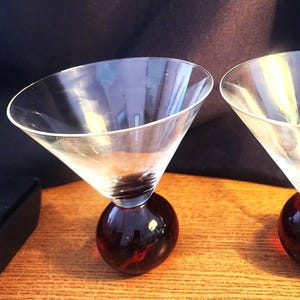 May include: Two clear glass martini glasses featuring wide, conical bowls and solid, spherical bases in a deep, translucent red color. The stems are absent, with the clear glass bowls resting directly atop the heavy, rounded red glass spheres.