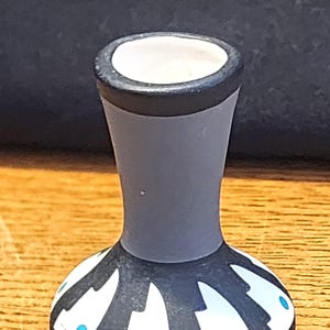 May include: A small, gray ceramic vase with a black rim and a decorative base. The base features a pattern of white shapes and turquoise dots. The vase has a narrow neck and a wide, rounded base. The interior is white.