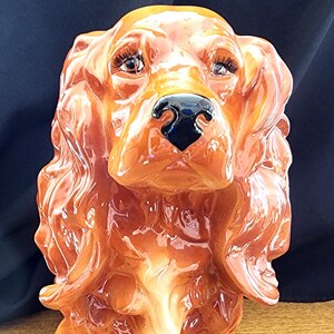 Puede incluir: Una figurita de cerámica de un cocker spaniel marrón y blanco. El perro está sentado con la cabeza inclinada hacia arriba y los ojos mirando hacia adelante. La figurita está esmaltada con un acabado brillante.