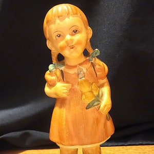 May include: A ceramic figurine of a young girl in a peach-coloured dress and brown shoes. She has blonde pigtails tied with blue ribbons and holds a bouquet of yellow flowers. The figurine is standing on a wooden surface.