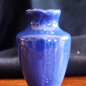 Puede incluir: Un pequeño jarrón de cerámica azul con una abertura ligeramente acampanada y un cuerpo redondeado. El jarrón tiene una superficie texturizada y moteada.