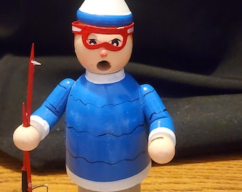 Puede incluir: Figura de madera de un esquiador con un suéter azul, gorro blanco y gafas y botas rojas. La figura sostiene bastones de esquí rojos y se para sobre una base blanca. El esquiador tiene una expresión de sorpresa.