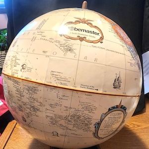 Może przedstawiać: Beżowy globus na brązowej drewnianej podstawie. Globus jest oznaczony jako "Globemaster" i ma średnicę 30,5 cm. Globus przedstawia kontynenty i oceany świata, ze szczególnym uwzględnieniem Oceanu Spokojnego. Globus zawiera również legendę wyjaśniającą różne ośrodki ludnościowe.