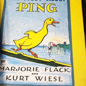 May include: A yellow hardcover book titled The Story About Ping by Marjorie Flack and Kurt Wiese. The cover features a yellow duck walking on a green plank over blue water, with a small boat and a pagoda visible in the background under a red sun.