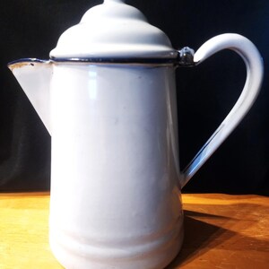 May include: White enamel coffee pot with a lid and a curved handle. The pot has a spout for pouring and a dark blue trim around the top and the lid. The pot is sitting on a wooden surface.