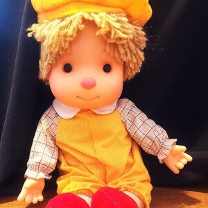 May include: A vintage doll with a yellow outfit, matching hat, and red shoes. The doll has blonde hair, a pink nose, and a white collar. The doll's arms and legs are outstretched.