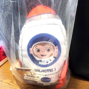 May include: A plush rocket ship toy in a clear plastic bag. The white rocket has red fins and an orange top. A cartoon monkey in a spacesuit is featured on the front, with the text "GALACTIC 1" and the NASA logo.