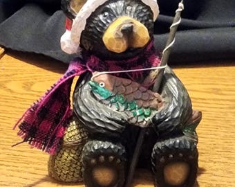 Puede incluir: Figura de oso de madera tallada con gorro de Papá Noel y bufanda a cuadros. El oso sostiene un pez y una caña de pescar. La figura tiene un aspecto rústico detallado, con vetas de madera visibles y detalles pintados.