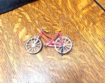May include: A miniature red bicycle with white wheels and yellow handlebars and seat. The small bike is resting on a wooden surface. The bicycle is a decorative item.
