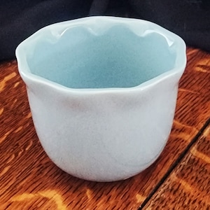 May include: A light blue ceramic cup with a scalloped rim. The cup is sitting on a wooden surface.