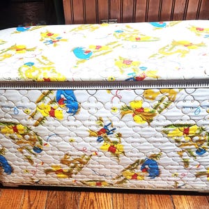 May include: A rectangular storage hamper with a white quilted vinyl exterior featuring a repeating pattern of yellow, blue, and red cartoon characters. The hamper has a matching lid and a white plastic trim along the top edge of the main body.