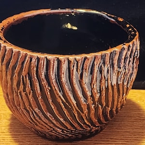 May include: A small, brown ceramic pot with a black glaze and a textured, wood-like pattern. The pot is empty and has a rounded shape.