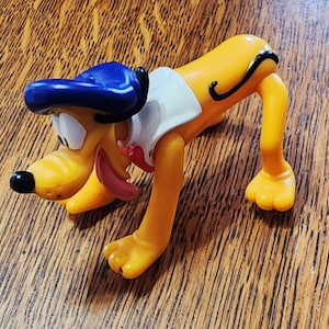 Könnte beinhalten: Ein gelber Plastik-Spielzeug-Hund mit einem blauen Hut und einem weißen Hemd. Der Hund ist in einer verspielten Pose mit seinen Beinen gebeugt und seinem Schwanz eingerollt.