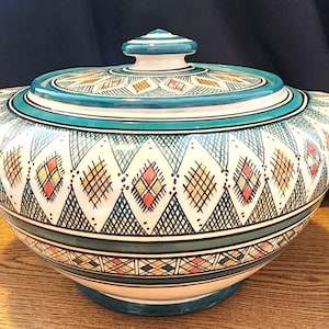 May include: A ceramic serving dish with a lid, featuring a white base and intricate geometric patterns in teal, yellow, red, and black. The dish has two handles and a teal rim, with a matching knob on the lid. The design is symmetrical and detailed.